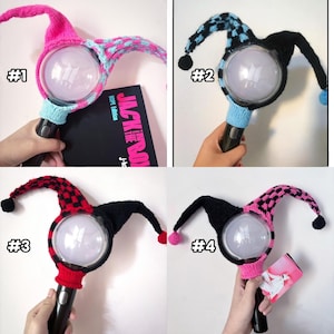 May include: Four light-up fan accessories with knitted covers in various color combinations. The covers feature jester-style designs with pointed tips and pom-poms. Each accessory has a black handle and a clear, round light. One image shows a book with the text "JACK IN THE BOX".