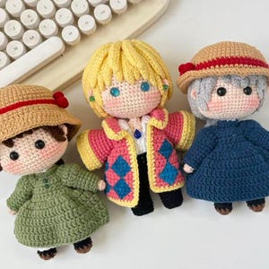 Puede incluir: Tres muñecas de ganchillo hechas a mano. Las muñecas tienen detalles, incluyendo sombreros, ropa y expresiones faciales. La muñeca de la izquierda lleva un vestido verde y un sombrero marrón con una banda roja. La muñeca central tiene un peinado amarillo y una chaqueta rosa. La muñeca de la derecha lleva un vestido azul y un sombrero marrón.