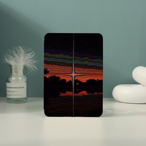 May include: A rectangular tablet case featuring a dark, symmetrical graphic design of a sunset over a silhouetted landscape with a central vertical light beam. The artwork displays wavy horizontal lines in shades of orange, red, and blue.