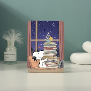 May include: A rectangular protective case featuring a cartoon illustration of a white dog resting against a pillow next to a tall stack of books. A small yellow bird sits atop the books reading. The background shows a night sky with stars.