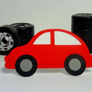May include: A red toy car with black wheels and a white roof. The car is sitting in front of a stack of black tires.