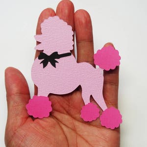 May include: Pink paper cutout of a poodle with a black bow. The poodle has pink pom-poms on its legs and tail.