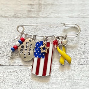 Pode incluir: Um alfinete de segurança prateado com pingentes, incluindo um coração com o texto "My heart belongs to a HERO", uma pequena bandeira americana, uma fita amarela e um pingente de contas vermelhas e azuis.