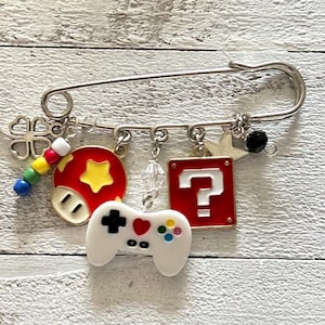 Op de afbeelding: Een zilveren veiligheidsspeld met bedels, waaronder een rode paddenstoel met een gele ster, een vraagtekenblok, een witte videogamecontroller, een klavertjevier en gekleurde kralen.