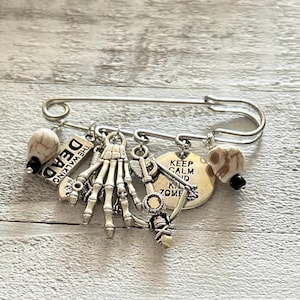 Könnte beinhalten: Eine silberne Sicherheitsnadel mit daran befestigten Anhängern. Die Anhänger umfassen einen Schädel, eine Skeletthand, einen Pfeil und einen runden Anhänger mit der Aufschrift "Keep Calm and Kill Zombies". Außerdem gibt es einen Anhänger mit der Aufschrift "The Walking Dead".