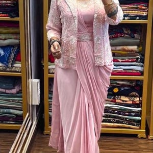 Puede incluir: Vestido formal rosa claro con chaqueta a juego. El vestido presenta un diseño drapeado y un cinturón enjoyado. La chaqueta está adornada con un delicado patrón bordado. El conjunto se exhibe en una tienda de ropa.