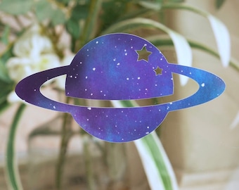Könnte beinhalten: Ein ausgeschnittener Papierplanet mit einem Ringsystem und zwei Sternen in Lila und Blau, im Galaxie-Design.
