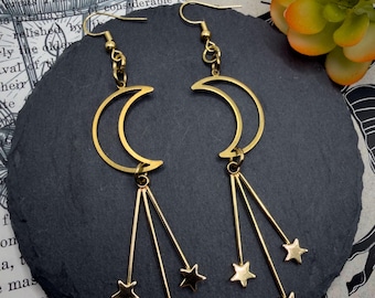 May include: A pair of gold-toned dangle earrings featuring a crescent moon charm at the top, with three thin metal rods extending downward, each ending in a small five-pointed star charm. The earrings are attached to standard gold-toned ear hooks.