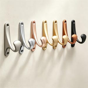 May include: A row of seven wall-mounted metal coat hooks arranged side by side. The hooks feature a curved, double-pronged design with two screw holes for mounting. The finishes include brushed nickel, polished chrome, rose gold, gold, brass, copper, and matte black.