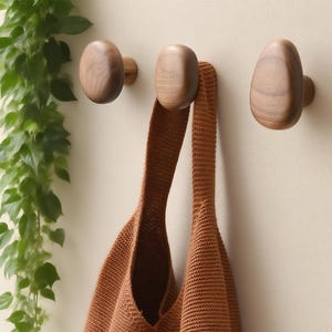 May include: Three wooden wall hooks with an oval design are mounted on a light-colored wall. A brown knitted tote bag hangs from one of the hooks. Green leafy vines are visible on the left side of the image.
