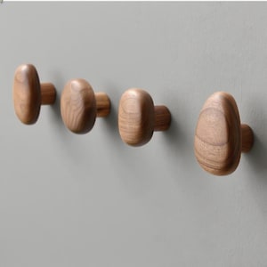 Puede incluir: Cuatro perchas de pared de madera con diseño ovalado. Los ganchos son de madera marrón claro y están montados en una pared gris. Los ganchos están espaciados uniformemente y son adecuados para colgar objetos. Cada gancho mide unos 5 cm de largo.