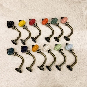 Può includere: Un set di dodici ganci da parete decorativi dall'estetica vintage. Ogni gancio presenta un braccio in metallo curvo color bronzo e un pomello in ceramica in vari colori, tra cui nero, giallo, rosso, arancione, verde e rosa.