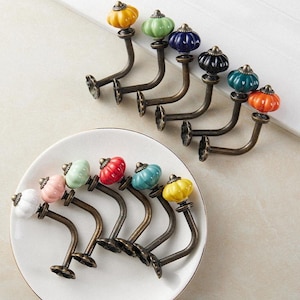 Puede incluir: Un conjunto de ganchos de pared decorativos con pomos de cerámica en forma de calabaza de varios colores. Los ganchos tienen un acabado de bronce y están dispuestos sobre una superficie blanca. Los colores incluyen amarillo, verde, azul y naranja.