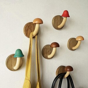 Könnte beinhalten: Wandhaken in Pilzform aus Holz in verschiedenen Farben, darunter Rot, Braun und Türkis. Die Haken sind auf runden Holzsockeln befestigt und halten einen gelben Schal und eine schwarze Handtasche. Das Design ist verspielt und dekorativ.