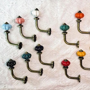Könnte beinhalten: Eine Sammlung dekorativer Wandhaken mit bunten, kürbisförmigen Keramikknöpfen. Die Haken haben eine Bronzefinish und sind vor einem weißen, strukturierten Hintergrund angeordnet. Farben sind Blau, Rosa, Weiß, Grün, Orange, Gelb, Rot und Schwarz.