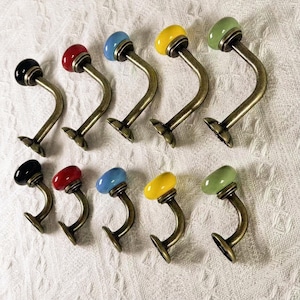 Puede incluir: Una colección de diez ganchos de pared decorativos con un acabado de latón vintage. Cada gancho presenta un brazo curvo y un pomo de cerámica redondeado y colorido en negro, rojo, azul, amarillo y verde. Los ganchos están dispuestos sobre una superficie blanca texturizada.