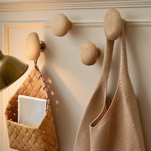 May include: Three wooden wall hooks with a natural finish are mounted on a light-coloured wall. A woven basket and a beige tote bag hang from two of the hooks. The hooks have a simple, rounded design, and the basket holds a few white cards.