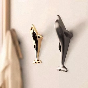 Peut inclure: Deux crochets muraux en forme de dauphin. L'un est de couleur or et l'autre est gris foncé. Les crochets sont montés sur un mur de couleur claire. Les crochets sont en métal et ont une surface lisse et réfléchissante.