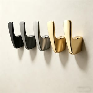 Könnte beinhalten: Vier moderne Wandhaken in einer Reihe, jeder mit einer anderen metallischen Oberfläche. Die Farben sind Schwarz, Dunkelgrau, Silber und Gold. Die Haken sind an einer hellbeigen Wand befestigt.