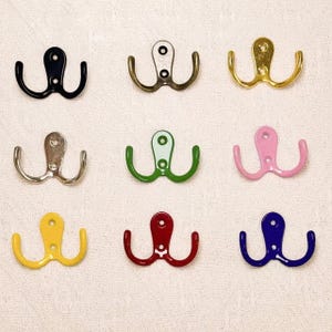 May include: Nine double coat hooks in various colors, including black, gold, silver, green, pink, yellow, red, and blue. Each hook has a rounded top with two curved hooks below, perfect for hanging coats or other items.