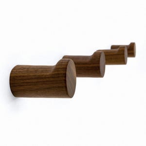 Könnte beinhalten: Vier hölzerne Wandhaken in einer Reihe, jeder mit einem zylindrischen Körper und einer gebogenen Oberseite. Die Haken sind aus dunkelbraunem Holz gefertigt und an einer weißen Wand montiert. Das Design ist minimalistisch und modern.