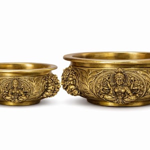 Puede incluir: Dos cuencos decorativos dorados. Los cuencos tienen un diseño ancho y poco profundo con un patrón de relieve detallado en los lados. El relieve representa figuras y elementos florales. Los cuencos son probablemente para uso decorativo.