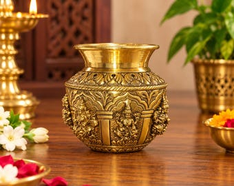 May include: A round brass pot featuring intricate embossed carvings of deities around its exterior. The vessel has a wide, flared rim and a polished metallic finish, showcasing detailed craftsmanship with repeating patterns and sculpted figures.