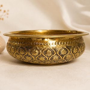 May include: A round, polished brass bowl featuring an intricate, repeating floral pattern embossed across its exterior surface. The bowl has a wide, smooth rim and a textured, darkened background within the floral design, creating a contrast.