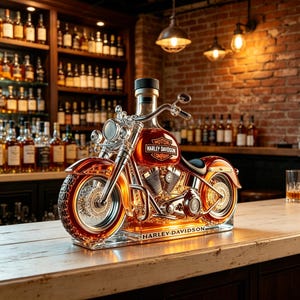 May include: A glass decanter shaped like a motorcycle with amber liquid inside. The fuel tank features the text MOTOR HARLEY-DAVIDSON COMPANY, and the base displays the text HARLEY-DAVIDSON. It has a black cap, chrome-colored details, and wheels.