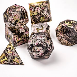 May include: A set of seven polyhedral dice for tabletop games. The dice are clear with a black and pink floral pattern and gold numbering. The dice include a d4, d6, d8, d10, d12, d20, and a percentile die.