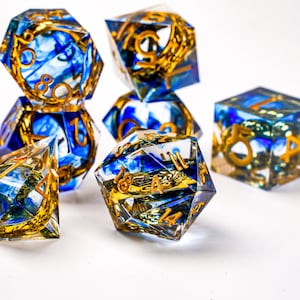 May include: A collection of polyhedral dice with a clear resin base, featuring blue and gold accents. The dice have gold numbering and intricate designs, suitable for tabletop gaming. The set includes various shapes, such as d4, d6, d8, d10, d12, and d20.