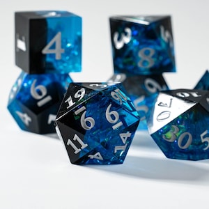 May include: A collection of polyhedral dice with a striking blue and black design. The dice feature a clear, resin-like material with embedded blue and silver accents. Each die has white numerals. The set includes various shapes, perfect for tabletop gaming.