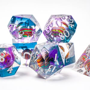 Puede incluir: Un conjunto de dados poliédricos con exteriores de resina transparente. Cada dado presenta una esfera colorida y arremolinada en su interior. Los dados tienen numeración plateada y son en tonos de azul, morado y transparente.