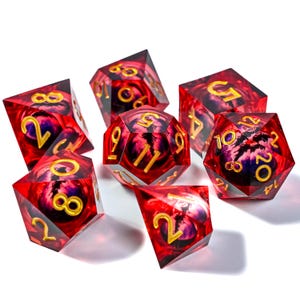 May include: Set of seven polyhedral dice. The dice are a transparent red with a swirling purple and black design inside. Gold numbers are visible on each face. The dice are for tabletop games.