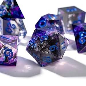 Puede incluir: Un conjunto de dados poliédricos con un diseño veteado en morado y negro. Los dados tienen números azules y están hechos de resina transparente. Para juegos de rol.