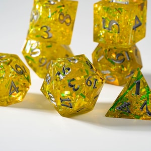 Könnte beinhalten: Eine Sammlung gelber, vielseitiger Würfel mit silbernen Zahlen. Die Würfel sind transparent und mit Glitzer gefüllt, was einen funkelnden Effekt erzeugt. Die Würfel sind auf einer weißen Oberfläche angeordnet, für Tischspiele.