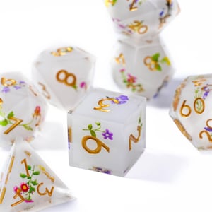 Op de afbeelding: Een verzameling witte dobbelstenen met gouden cijfers en bloemmotieven. De dobbelstenen hebben verschillende vormen, waaronder een kubus, een piramide en een veelzijdige dobbelsteen. De bloemmotieven bevatten paarse, roze en groene accenten.
