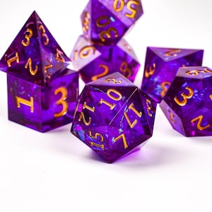 Puede incluir: Juego de siete dados poliédricos en morado transparente con numeración dorada. Los dados tienen varias formas, incluyendo un d4, d6, d8, d10, d12, d20 y un dado de porcentaje. Los dados están dispersos sobre una superficie blanca.