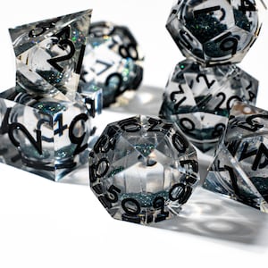 May include: A collection of clear resin dice with black numbering and embedded glitter. The dice are various polyhedral shapes, including a d4, d6, d8, d10, d12, and d20. The glitter is a dark teal color, adding a shimmering effect.