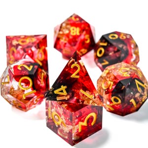 May include: Set of six polyhedral dice in various shapes, featuring a red and gold color scheme. The dice have gold numbering and are designed for tabletop role-playing games. The dice are made of a clear resin with gold flakes.