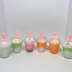 May include: Six colorful bubble tea keychains with clear plastic cups, each topped with whipped cream and a pink straw. The drinks are various colors, including green, pink, and orange, and have silver key rings.