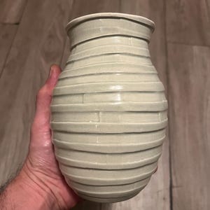 May include: A light gray ceramic vase with a rounded, bulbous body and a narrow neck. The exterior surface features a series of horizontal, raised ridges that wrap around the entire circumference of the vessel, creating a textured, layered appearance.