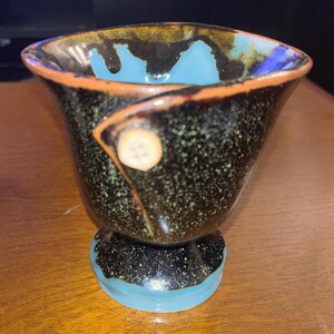 May include: A handmade ceramic vase with a unique design. The vase features a speckled black exterior with a turquoise interior and base. An orange rim and a decorative button add to its artistic appeal. The vase is approximately 4 inches tall.