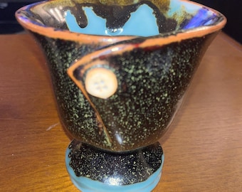 Puede incluir: Un jarrón de cerámica hecho a mano con un diseño único. El jarrón presenta un exterior negro moteado con un interior y base turquesa. Un borde naranja y un botón decorativo se suman a su atractivo artístico. El jarrón mide aproximadamente 10 cm de alto.
