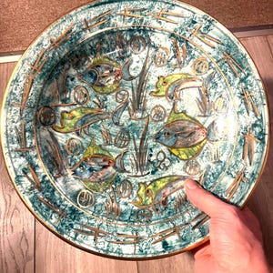 Puede incluir: Un plato decorativo de cerámica con un diseño turquesa y dorado. El plato presenta peces y plantas acuáticas pintados a mano. El borde está adornado con oro. El plato es redondo y tiene un acabado brillante.