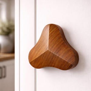 Puede incluir: Un pomo de puerta de madera con un diseño geométrico único. El pomo es de color marrón medio cálido y está montado en una puerta blanca. El diseño presenta una forma triangular con bordes redondeados, añadiendo un toque moderno.