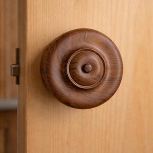 May include: A close-up of a wooden cabinet knob with a circular design. The knob is a dark brown color and has concentric circles carved into its surface. The knob is mounted on a light-colored wooden cabinet door.