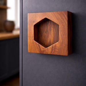 May include: A square, wooden cabinet pull with a hexagonal cutout. The pull is made of a warm-toned wood and is mounted on a dark gray cabinet door. The wood grain is visible.