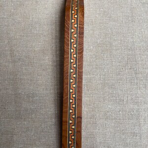 May include: A long, rectangular wooden bookmark with a decorative inlaid design. The bookmark is primarily brown with a central band of orange, cream, and black geometric patterns. The bookmark is approximately 10 inches long.