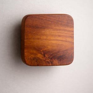 May include: A square wooden box with rounded edges. The box is made of a rich, warm-toned wood, showcasing the natural grain patterns. The design is simple and minimalist, with a focus on the wood's texture and color.